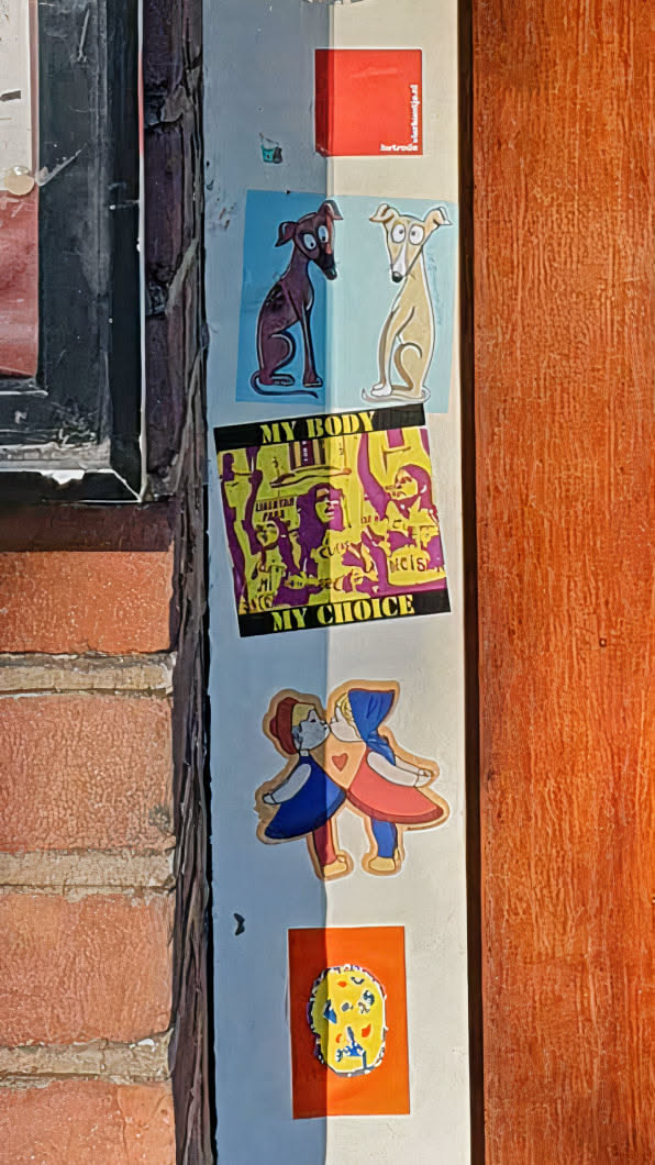 A series of stickers are affixed to a wall, showcasing a variety of designs. One features two greyhounds, one brown and one beige, against a light blue background. Below this is a "My Body My Choice" sticker with figures protesting in purple and yellow. Another sticker depicts two figures in blue and red kissing with a heart between them. The stickers are arranged vertically.
