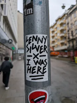 A rectangular, vertically oriented sticker with a white background and bold, black, handwritten-style text. The text reads 'LENNY SILVER' at the top, followed by a horizontal line with an 'X' in the middle, and then 'I WAS HERE' at the bottom, also underlined by a horizontal line. The sticker appears slightly wet with visible water droplets, indicating it's exposed to the elements. Its edges show minor wear, suggesting it's a physical sticker adhered to a metal pole.