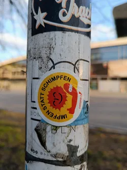 A round sticker with a yellow sun-like design and a figure holding a syringe. The text "Impfen statt Schimpfen!" is written around the design, suggesting a pro-vaccination message.  The sticker is affixed to a pole covered in other stickers and grime.