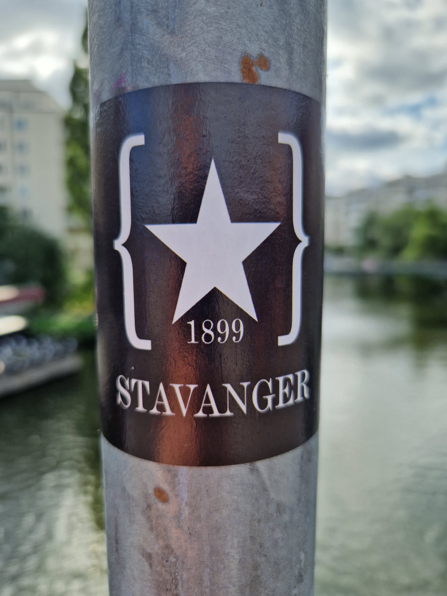 A dark-colored sticker with a white star and the year 1899, along with the word 'Stavanger' printed in a serif typeface.  The star is enclosed in stylized curly brackets. The sticker is affixed to a metal pole by a body of water.