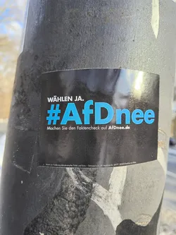 A rectangular sticker is affixed to a gray pole. The sticker has a black background and blue text that reads 'WÄHLEN JA. #AfDnee Machen Sie den Faktencheck auf AfDnee.de'