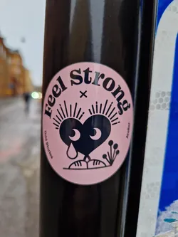 Pink and black round sticker with illustration of a crying heart character.  The text 'Feel Strong' is written in a circular fashion around the illustration. Small text at the bottom reads 'Odd stories' and 'Atelier'.