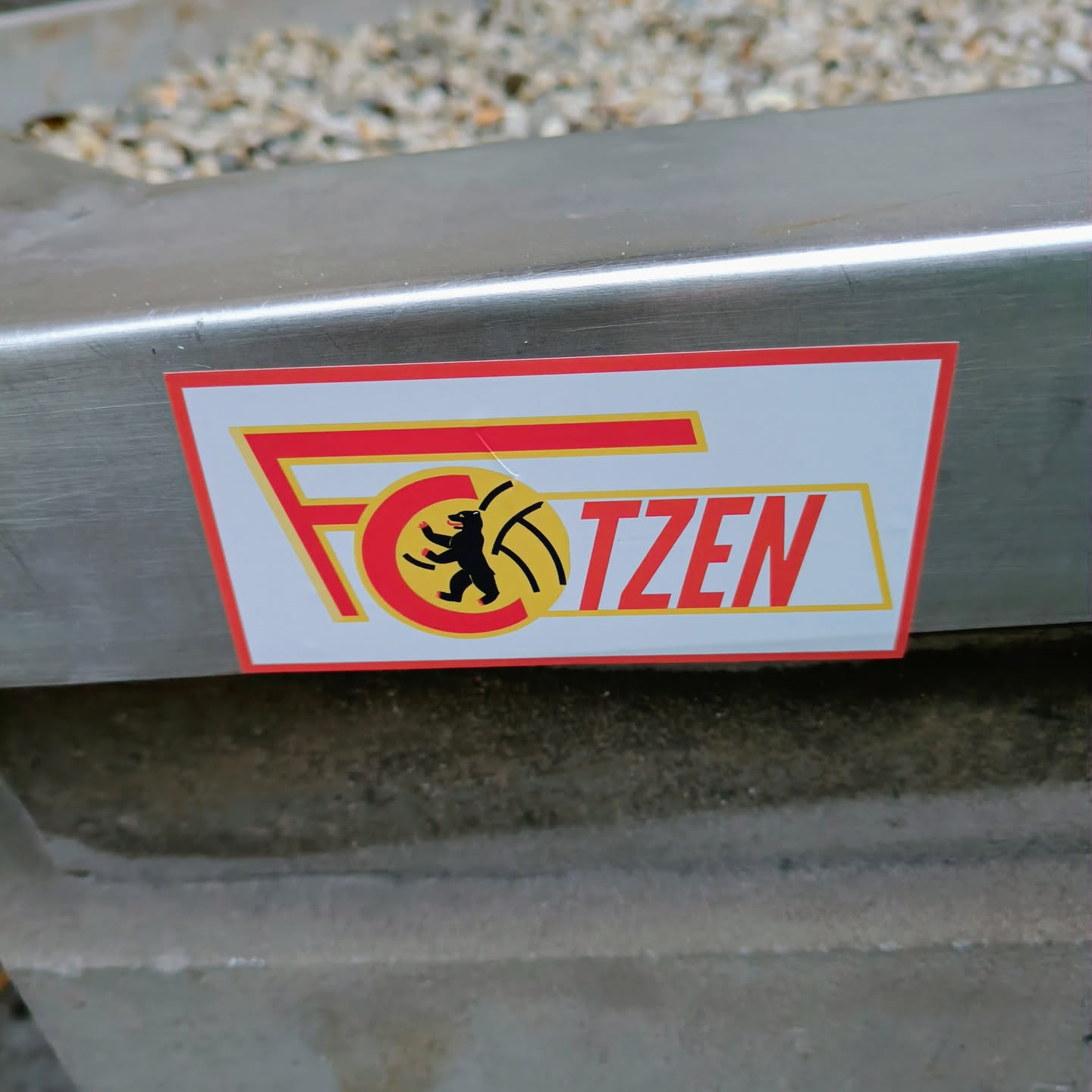A rectangular sticker featuring the logo of FC Kötzen. The logo has a red and yellow design with a bear emblem. The text 'FC' and 'KÖTZEN' is written in a bold font. The sticker is attached to a metal surface.