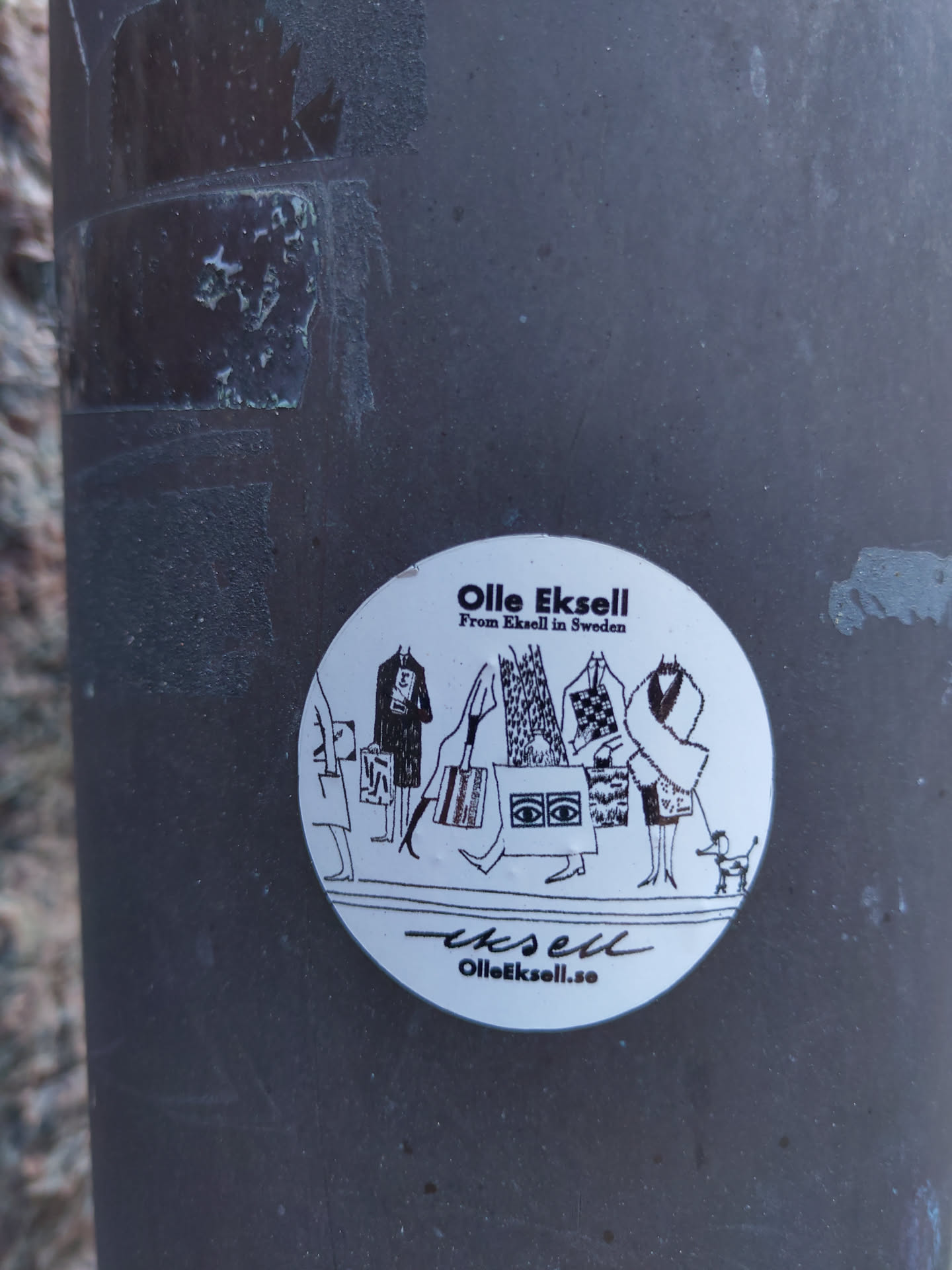 Round sticker featuring a black and white illustration of people shopping, carrying bags, and a small dog. The artist's name and website are included at the bottom.