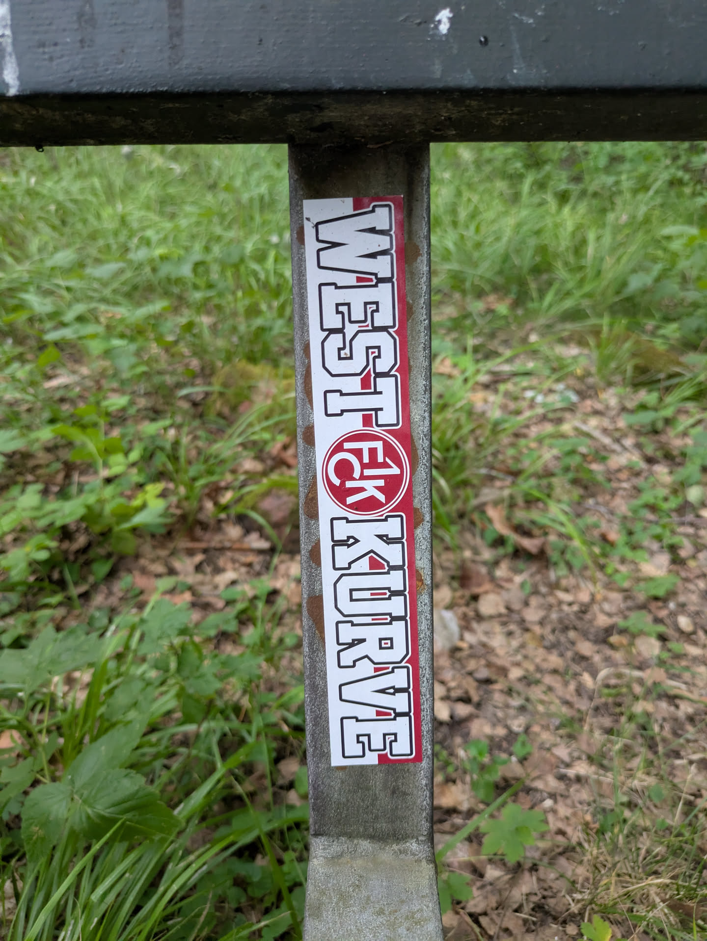 A white rectangular sticker with a red border, oriented vertically on a weathered grey metal pole. The sticker features the text "WEST KURVE" written in bold white block letters with a black outline, running from top to bottom. In the middle of the red section, there's a red circle containing the white letters "FCK", likely referring to a football club. The background is a mix of green grass and fallen leaves, suggesting an outdoor, natural setting, possibly near a sports ground or park.