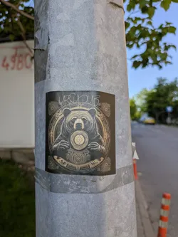 A square sticker adhered to a metal pole features a detailed illustration of a bear holding a record in its hands. The artwork has an intricate, almost ornamental quality. The text "URSUDUB" is visible above the bear.