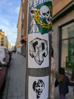 A black and white sticker featuring an abstract cube with a stylized letter or symbol inside. The style is reminiscent of a geometric puzzle or a 3D design. The sticker is placed on a pole with other stickers, suggesting street art or urban decor.