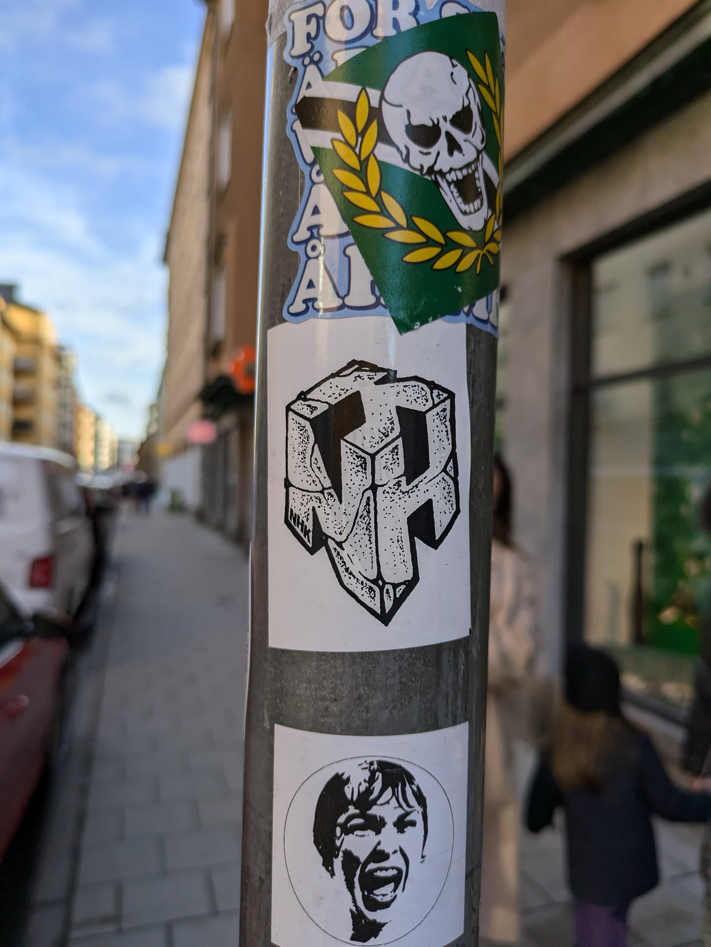 A black and white sticker featuring an abstract cube with a stylized letter or symbol inside. The style is reminiscent of a geometric puzzle or a 3D design. The sticker is placed on a pole with other stickers, suggesting street art or urban decor.