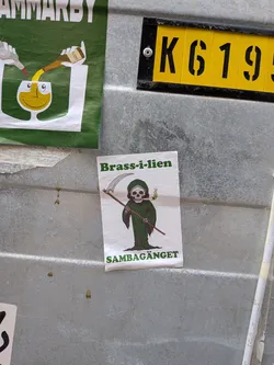 The sticker features a cartoonish depiction of the Grim Reaper. The Reaper has a white skull for a head with dark eye sockets and nose cavity. It wears a dark green hooded robe that covers its body, with tattered edges at the bottom. Its skeletal hands are visible, holding a long, light brown wooden scythe with a silver blade. Notably, the Reaper is smoking a lit cigarette or joint, which emits a small puff of green smoke. The background of the sticker is plain white. Above the Reaper, the text 'Brass-i-lien' is written in green, and below it, 'SAMBAGÄNGET' is also in green.