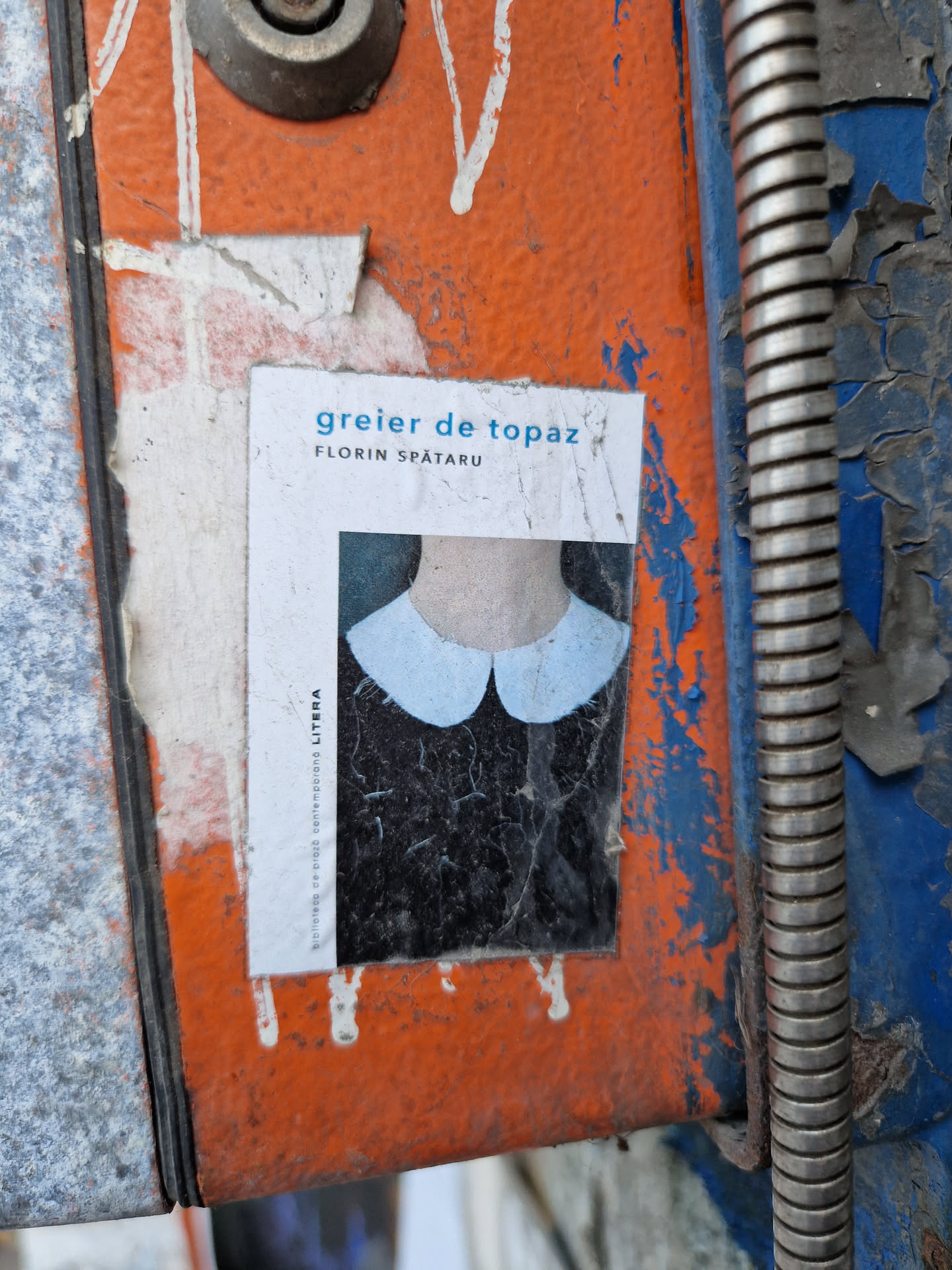 A sticker featuring the cover of a book titled "greier de topaz" by Florin Spătaru. The cover shows a person wearing a light blue Peter Pan collar. The sticker is affixed to a weathered, orange and blue metal surface.