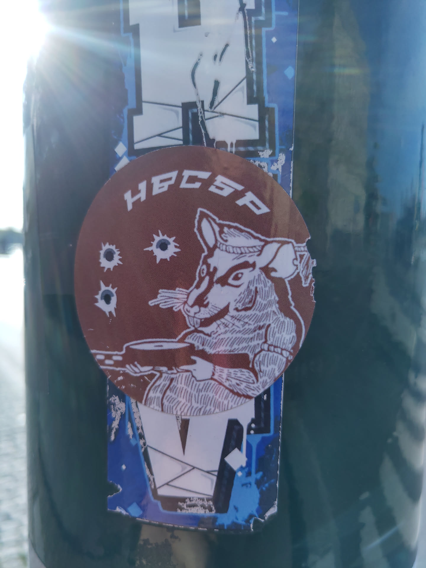 A round brown sticker with a drawing of a rat holding what looks like a weapon. There are three bullet holes visible on the sticker around the rat. The text HBCSP is written on top of the sticker. The sticker is placed on a dark green surface, with other stickers underneath and nearby. 