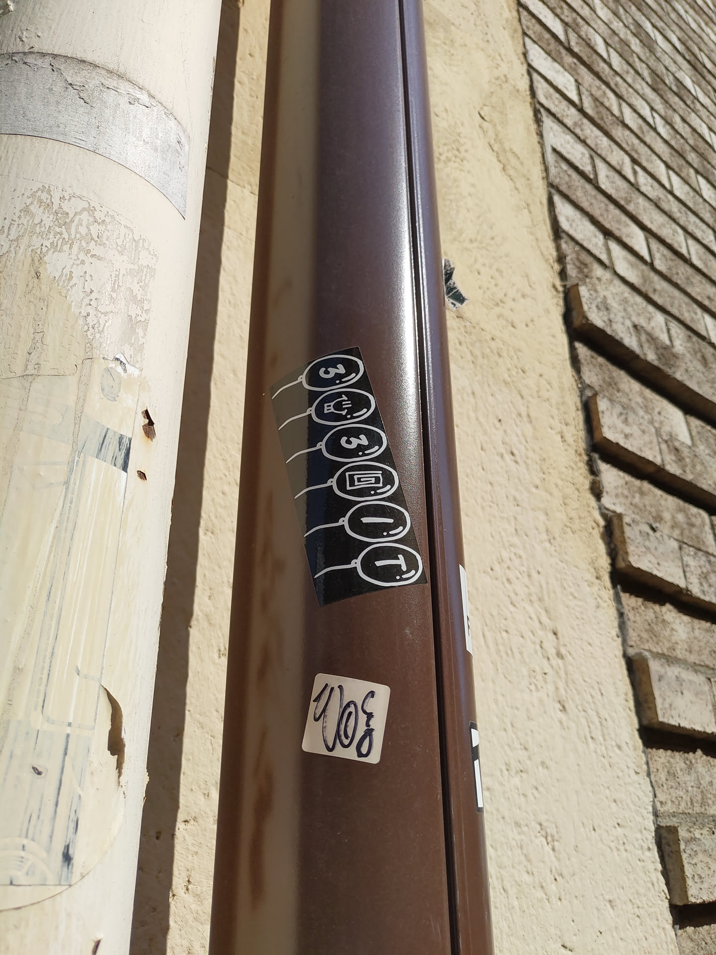 A vertical sticker featuring five circular icons connected by lines. The icons appear to be stylized symbols, possibly representing different functions or concepts. The sticker is affixed to a brown downspout on a building.  Another smaller sticker with illegible writing is below it.
