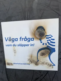 A sticker with a public safety message, the sticker warns to be aware of whom you let in. There is an illustration of a person in a striped shirt helping someone over a step. The sticker is partially burned.