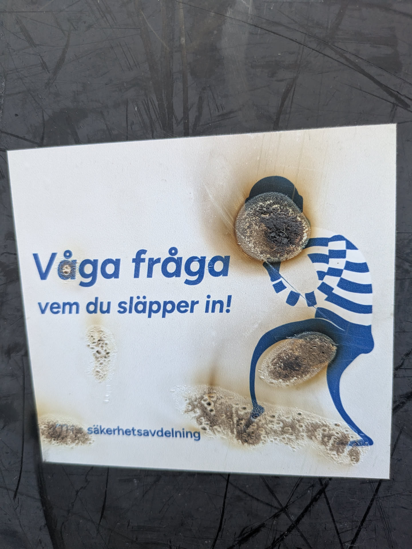 A sticker with a public safety message, the sticker warns to be aware of whom you let in. There is an illustration of a person in a striped shirt helping someone over a step. The sticker is partially burned.