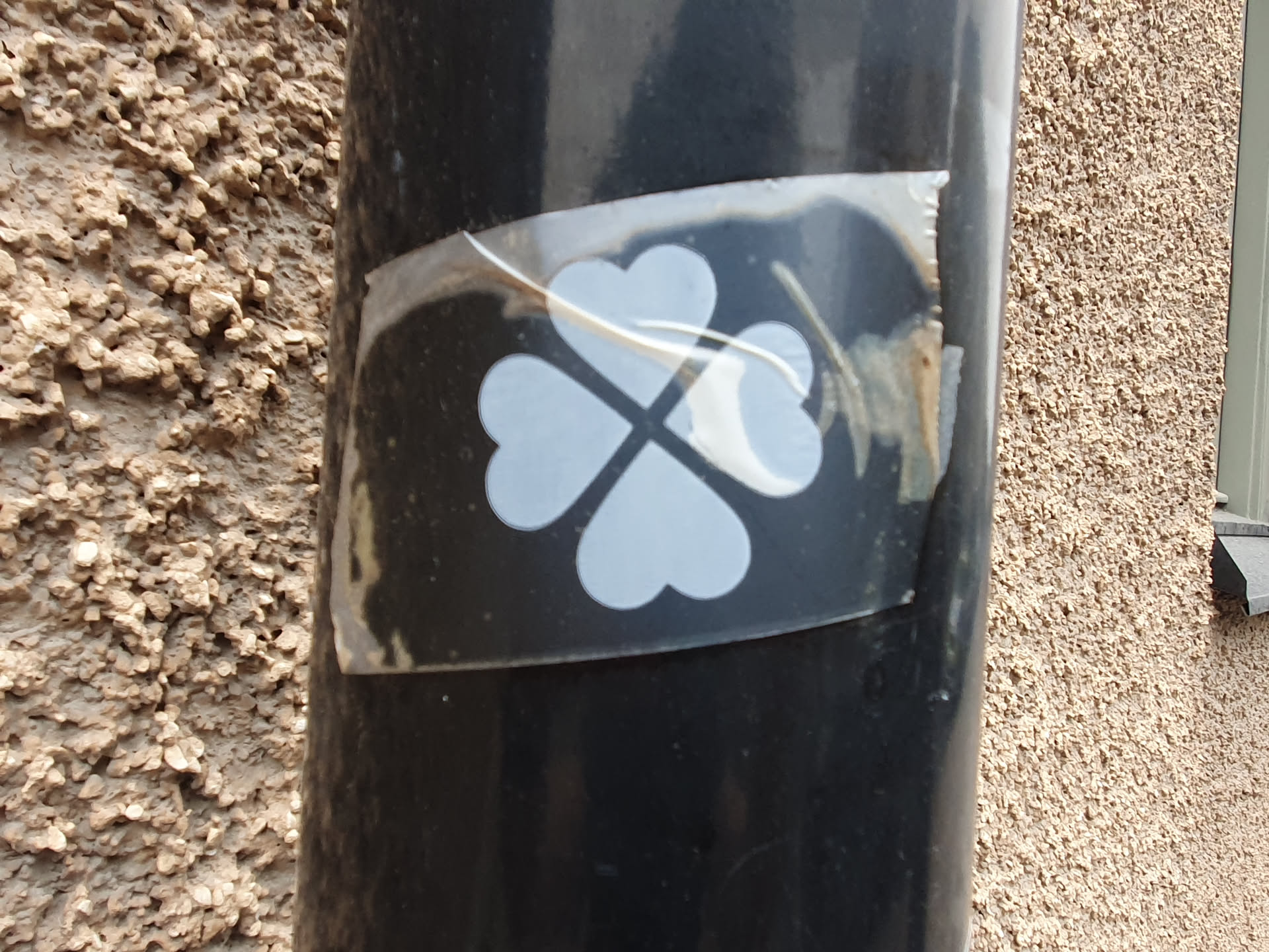 A black and white sticker featuring a four-leaf clover design. It's affixed to a dark-colored pole against a textured wall. The sticker appears slightly worn or damaged around the edges.