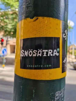 A black and yellow sticker with the word Snösåtra and their website address on it. The sticker is attached to a green pole, with a honeycomb-like pattern on the yellow background. 