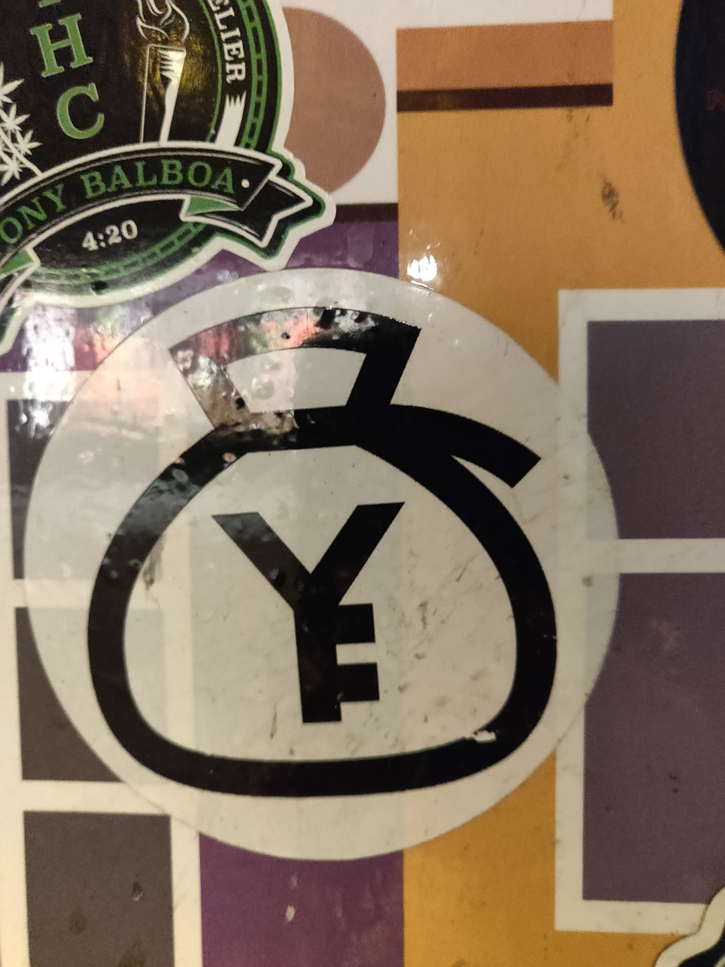 A round sticker featuring a stylized Yen symbol in black on a white background. The sticker shows some wear and tear.