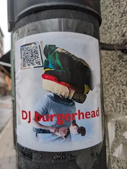 Sticker featuring an illustration of a person with a burger-shaped head, styled as a DJ.  A QR code is present in the top left corner. The style is colorful and slightly surreal.