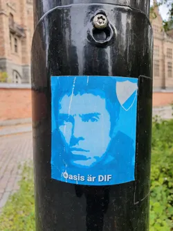 A blue sticker featuring a stylized portrait and the text "Oasis är DIF".  The sticker is affixed to a dark-colored pole outdoors.