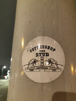 Round sticker with black and white illustration of two bulls facing each other with a coat of arms in between. The text on the sticker says "Coffeeshop The Stud", "Amsterdam", "Sinds 1982".