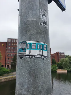 A sticker depicting a stylized teal and white subway train. The sticker is affixed to a grey metal pole outdoors near water.  Another sticker featuring a Batman-esque character is visible above it.