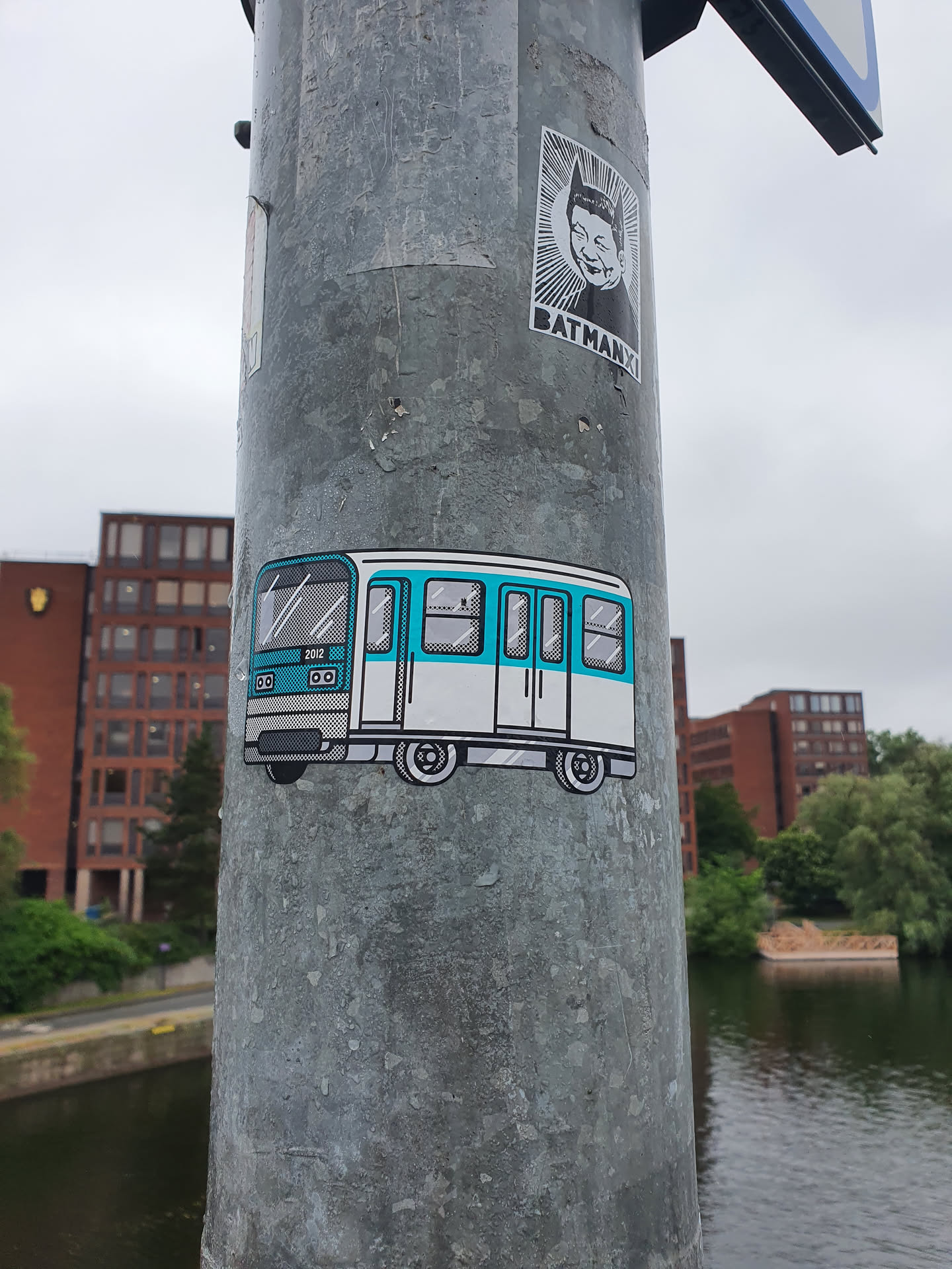 A sticker depicting a stylized teal and white subway train. The sticker is affixed to a grey metal pole outdoors near water.  Another sticker featuring a Batman-esque character is visible above it.