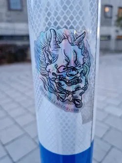 Holographic sticker featuring an illustration of an Oni mask. The sticker is affixed to a reflective pole. The bottom of the sticker contains a website address.