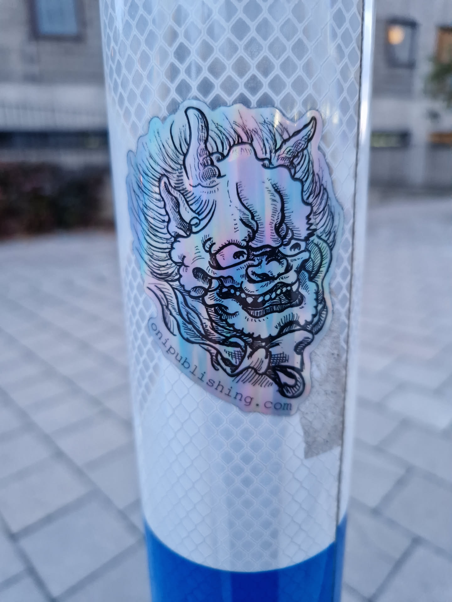 Holographic sticker featuring an illustration of an Oni mask. The sticker is affixed to a reflective pole. The bottom of the sticker contains a website address.