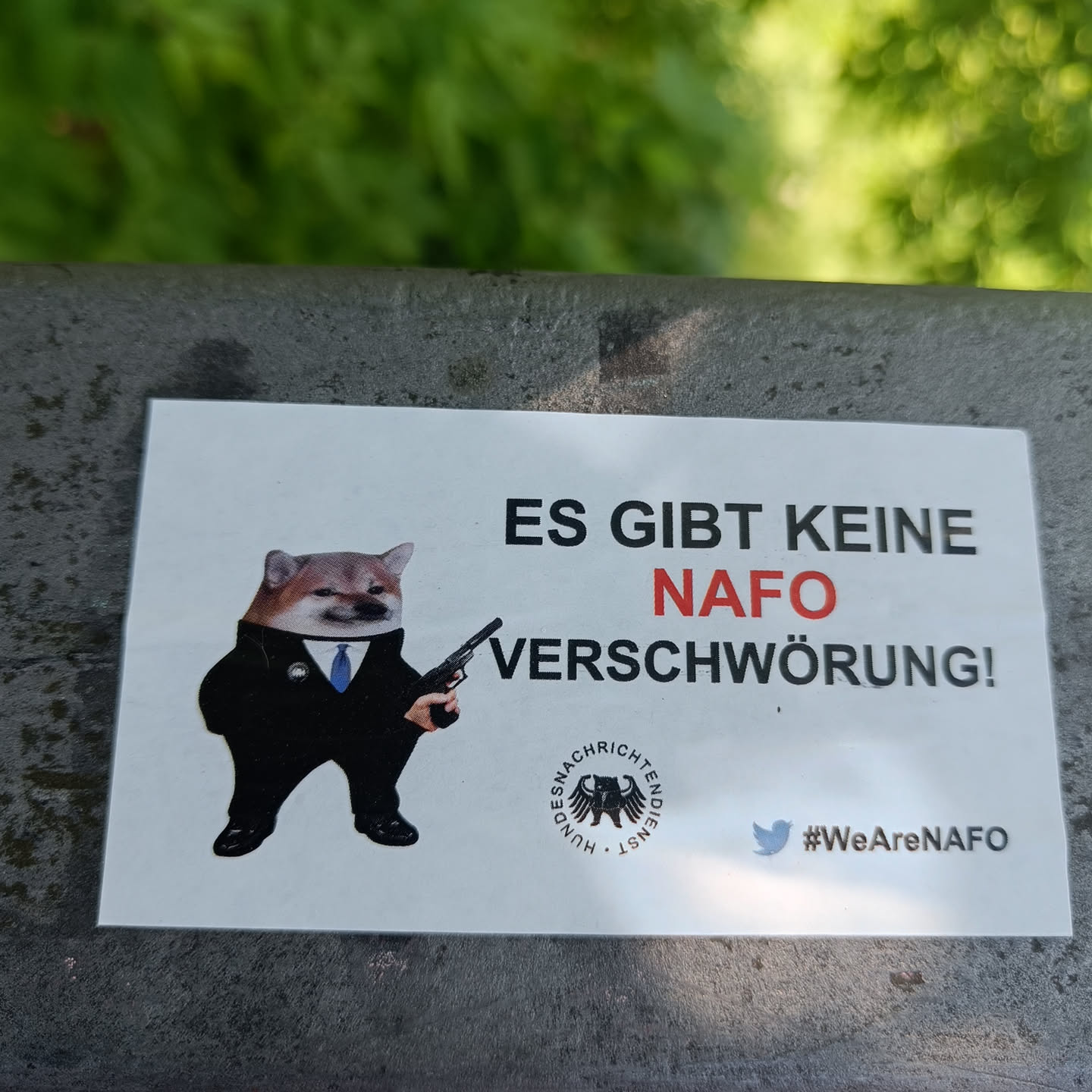 A sticker featuring a Shiba Inu in a suit holding a gun, with text in German stating "There is no NATO conspiracy!" and a Twitter hashtag #WeAreNATO.  The sticker also includes a logo that appears to be related to German news.