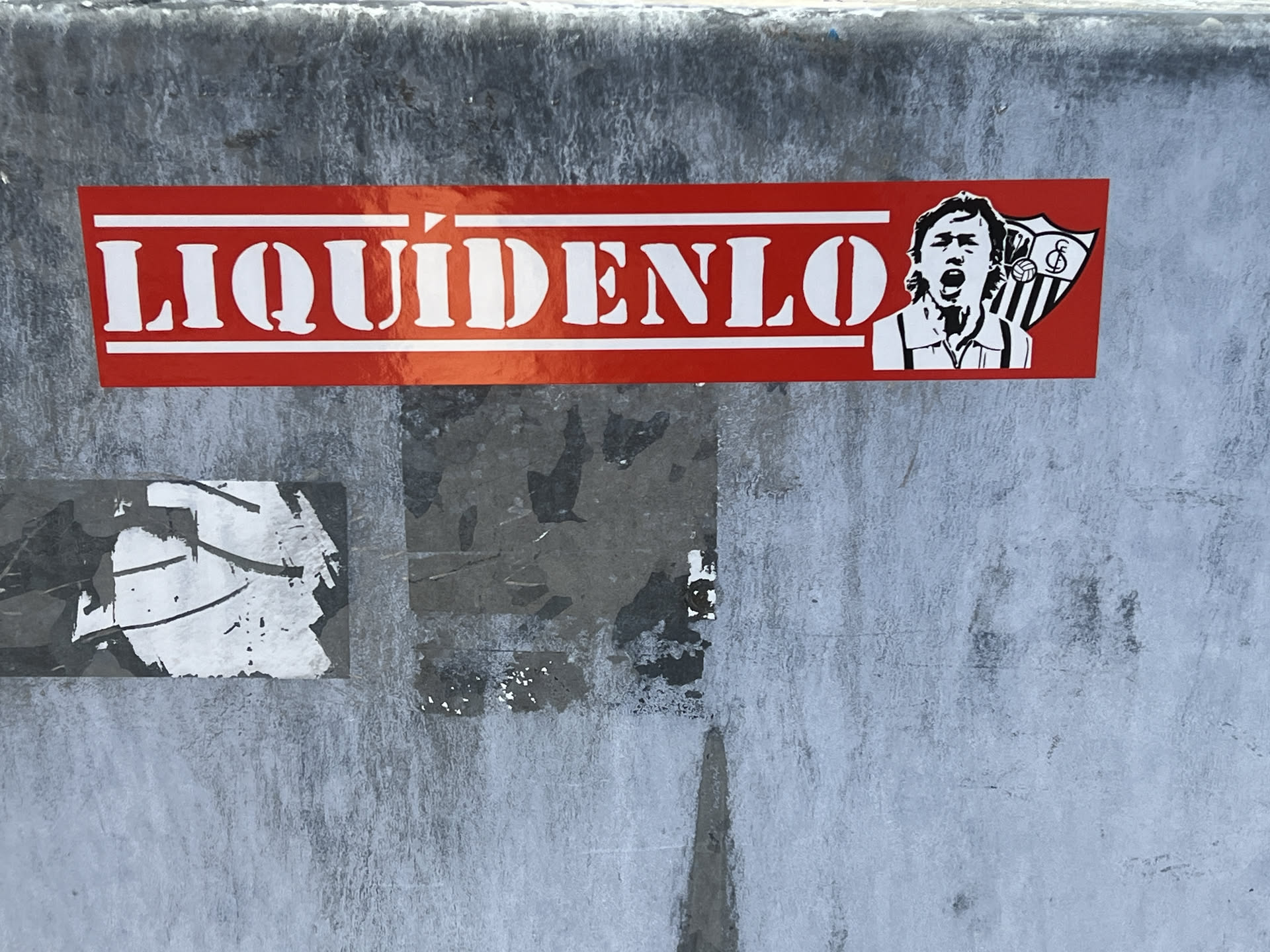 A rectangular sticker with a vibrant red background. The main text, 'LIQUÍDENLO', is prominently displayed in white, bold, serif-style letters with a slightly distressed or stencil-like appearance, framed by thin white lines above and below. To the right, there is a black and white illustration of a male figure, likely a football player, with an open mouth, appearing to shout or cheer. Behind this figure is the crest of Sevilla Fútbol Club, featuring vertical red and white stripes, a stylized 'SFC' monogram, and a football. The sticker has a glossy finish, showing some reflections, and is adhered to a textured, weathered grey surface.