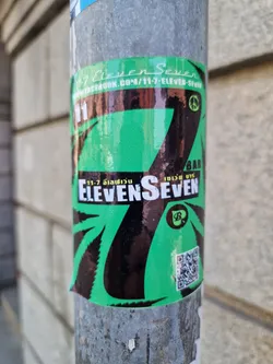 A green and black sticker with the number 7 prominently displayed.  The number is stylized and incorporates a cannabis leaf design.  The text "Eleven Seven" is visible, along with a peace symbol and what appears to be a logo or brand mark. A QR code is also present on the sticker.