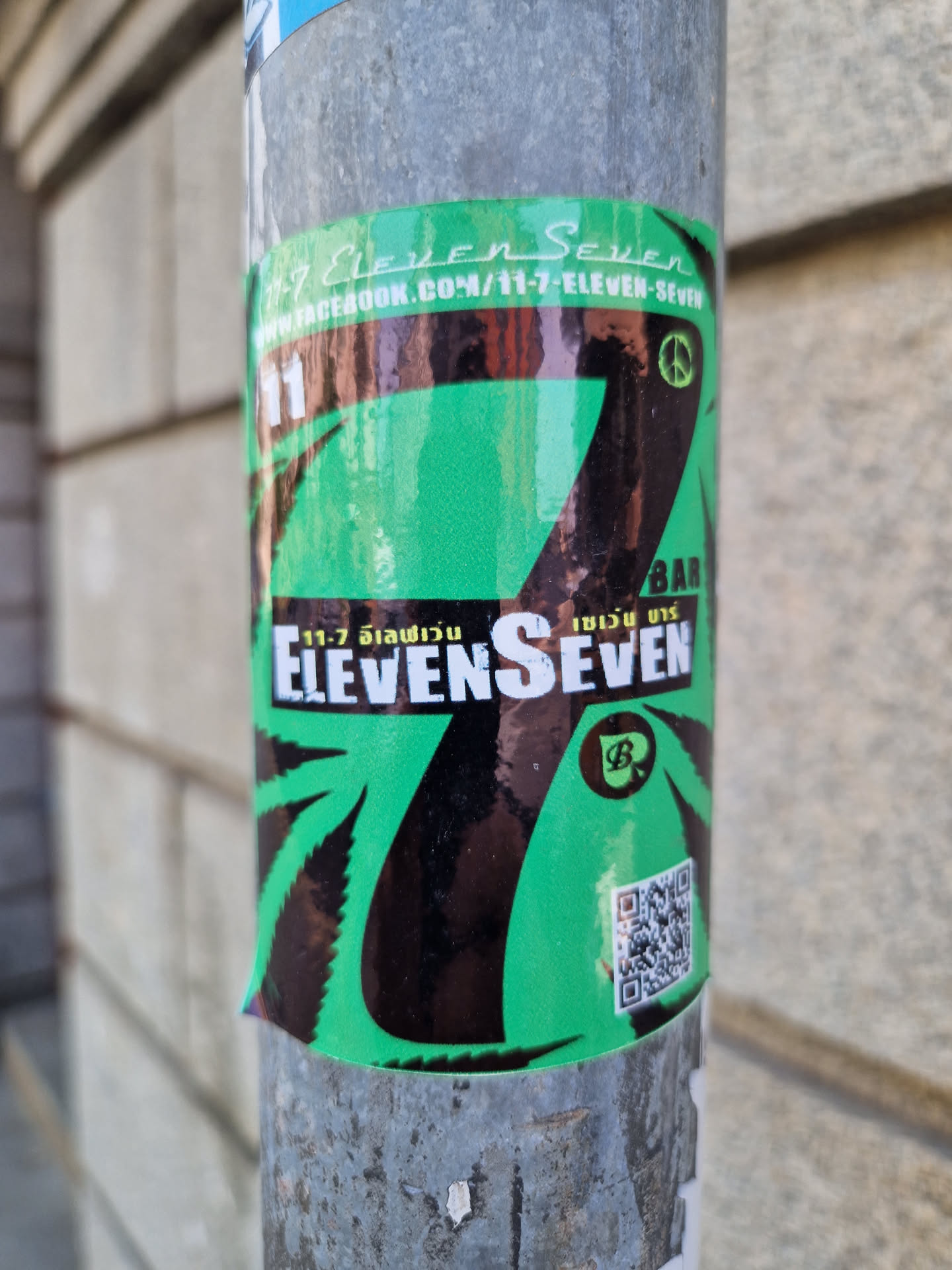 A green and black sticker with the number 7 prominently displayed.  The number is stylized and incorporates a cannabis leaf design.  The text "Eleven Seven" is visible, along with a peace symbol and what appears to be a logo or brand mark. A QR code is also present on the sticker.