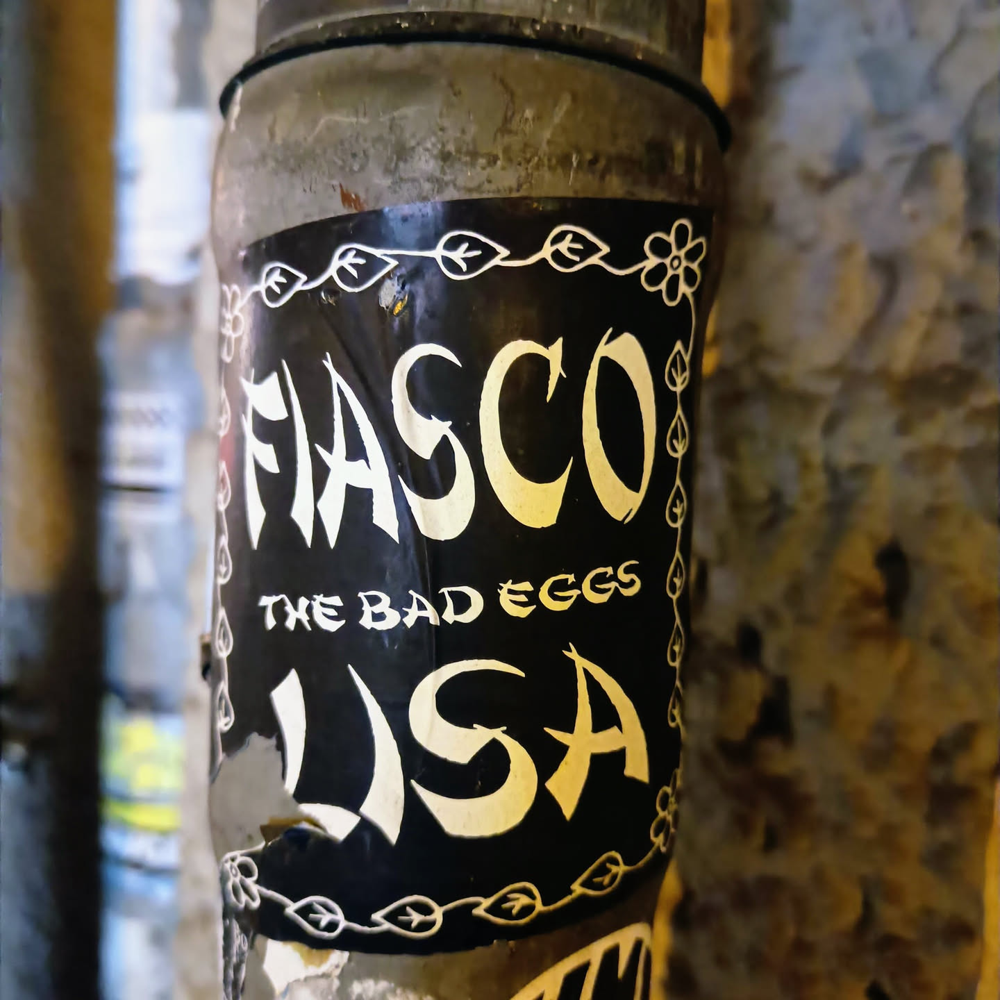 A black and white sticker with the text "FIASCO" in a stylized font, with the words "THE BAD EGGS" underneath and "USA" below that. The sticker has a decorative border of leaves and flowers. It is affixed to a weathered metal pipe.