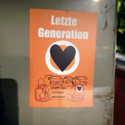 An orange sticker with a black heart in the center surrounded by a white circle. Below it, there is an illustration of people sitting with backpacks, and cars in the background. The text on the sticker says "Letzte Generation" at the top and "Stoppt den fossilen Wahnsinn" (Stop the fossil madness) at the bottom.