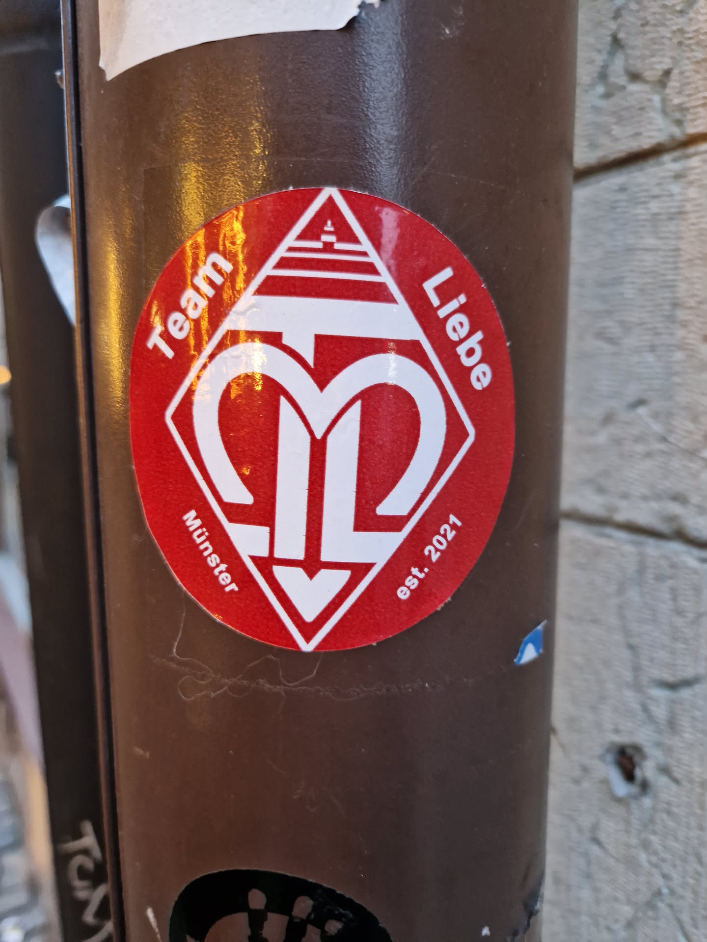 Red and white diamond-shaped sticker with a logo and text. The logo is a stylized "M" with an arrow pointing down. The text around the logo says "Team Liebe" at the top and "Münster est. 2021" at the bottom.