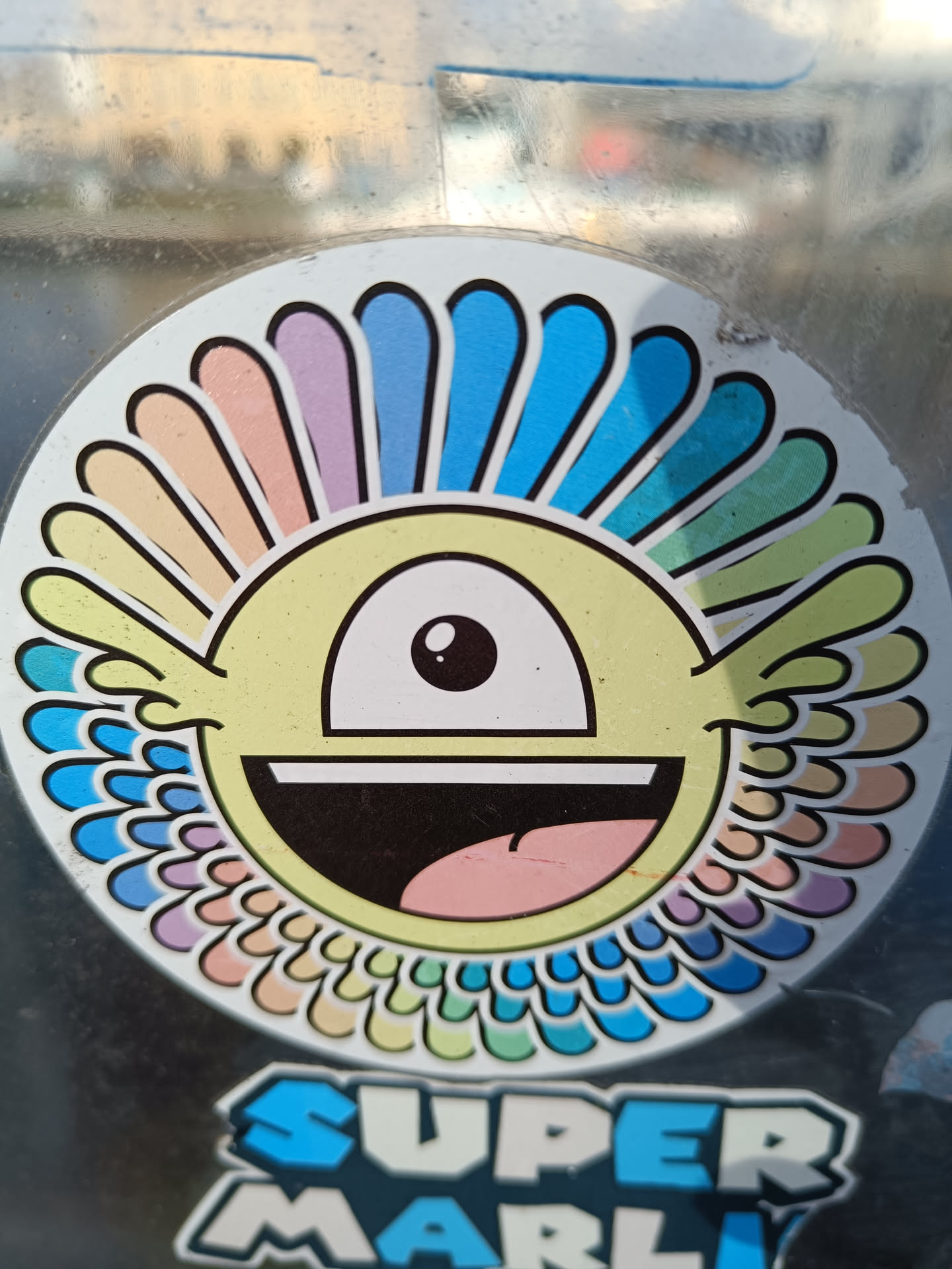 This sticker showcases a vibrant, cartoonish character with a single eye and a wide, joyful smile. Its design incorporates a captivating rainbow-colored halo around its head, creating a psychedelic and playful aesthetic. The text "Super Marly" is prominently displayed below the main character. 