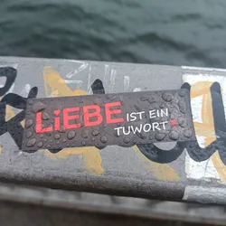 A dark gray rectangular sticker with the German phrase "Liebe ist ein Tuwort" (Love is a deed) in red and white lettering.  The sticker is affixed to a surface covered in other graffiti and is wet with water droplets.