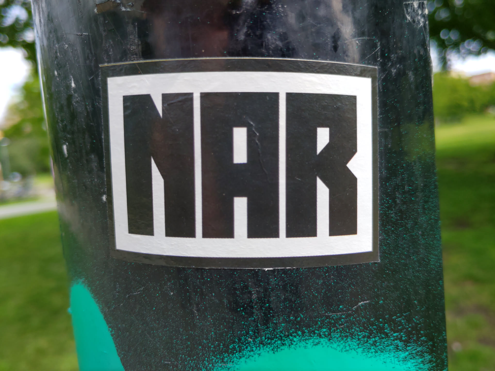 A black and white rectangular sticker with the letters NAR in bold, uppercase, sans-serif font. The sticker is affixed to a dark-colored pole outdoors. 