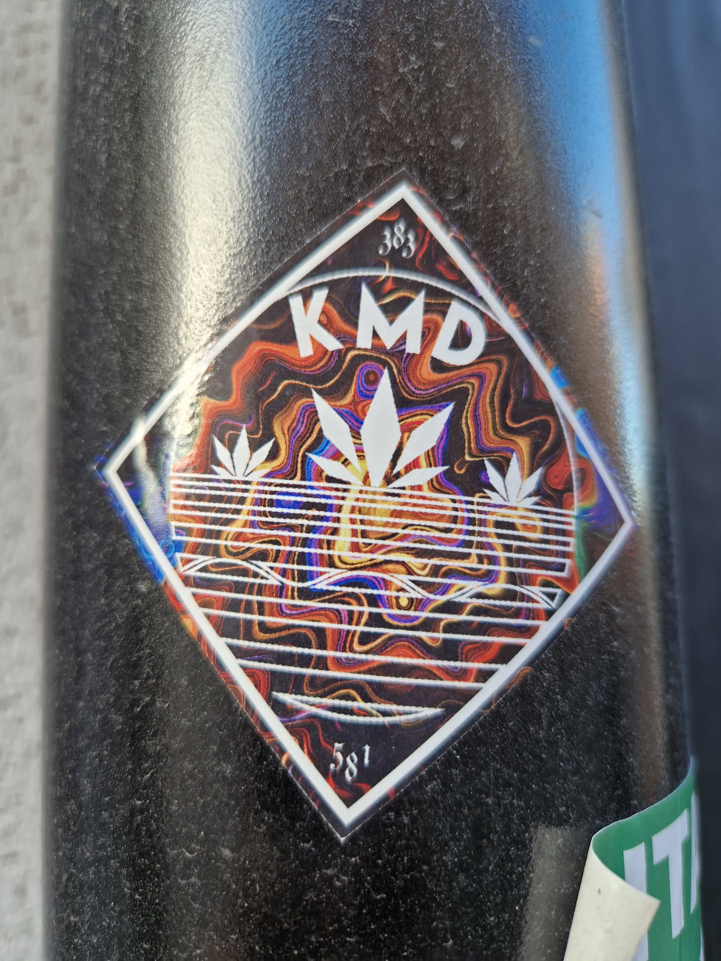 Diamond-shaped sticker featuring the acronym KMD in the center, surrounded by a psychedelic wave-like pattern with three cannabis leaves. The numbers 383 and 581 are visible at the top and bottom of the sticker, respectively. The overall color scheme is a mix of dark and light colors with a vibrant psychedelic theme.
