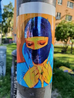 A vibrant sticker depicting a stylized portrait of a person in a blue balaclava. The artwork is rendered in a bold, graphic style with bright colors and a slightly surreal aesthetic. The background is a warm orange-yellow, contrasting with the cool blues and yellows of the figure. Some text is partially visible on the figure's clothing, but it's not fully legible.  The sticker appears to be affixed to a metal pole outdoors.