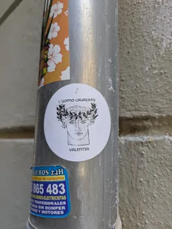 This sticker features a black and white drawing of a human face wearing a laurel wreath. The title above the drawing says "L'UOMO LAUREATO" and "VALENTIA" is written below the drawing. The sticker is round.