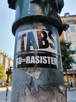 A sticker with the text "TABS" on top and "SD=RASISTER" (SD=Racist) on the bottom, affixed to a lamppost. The sticker displays a strong anti-racist message, likely targeting the Sweden Democrats political party.