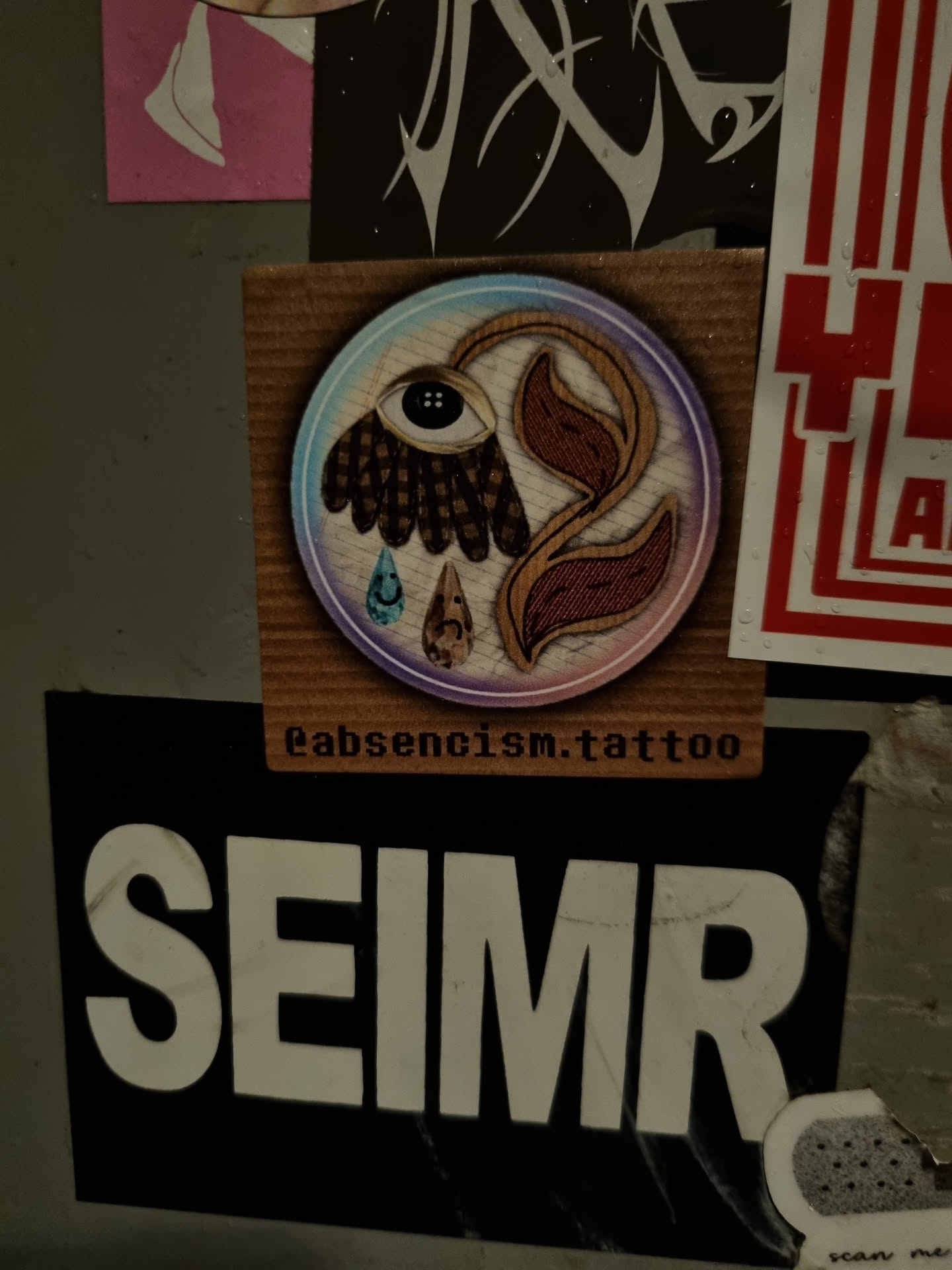 A round sticker with a detailed illustration of a stylized flower incorporating an eye and a single teardrop. The flower is depicted in earthy tones with hints of purple and blue. The text 'eabsentism.tattoo' is visible beneath the main illustration.