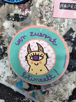 This round sticker showcases a cute, yellow monster with a single, large purple eye. The creature has small horns and a charming smile, set against a background pattern of numerous tiny eyes. The text 'Wer zuerst blinzelt!' curves around the design, written in a playful, handwritten style.