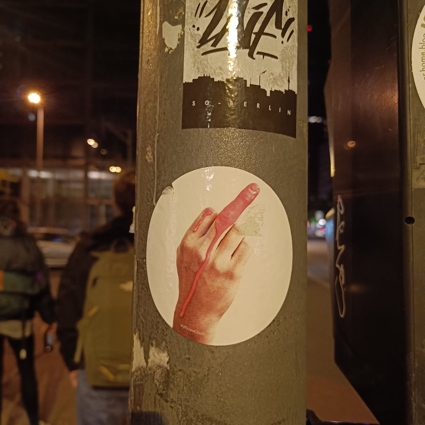 A round sticker featuring a hand giving the middle finger. The sticker is affixed to a pole in what appears to be an urban setting. The background is blurred, suggesting a night scene.