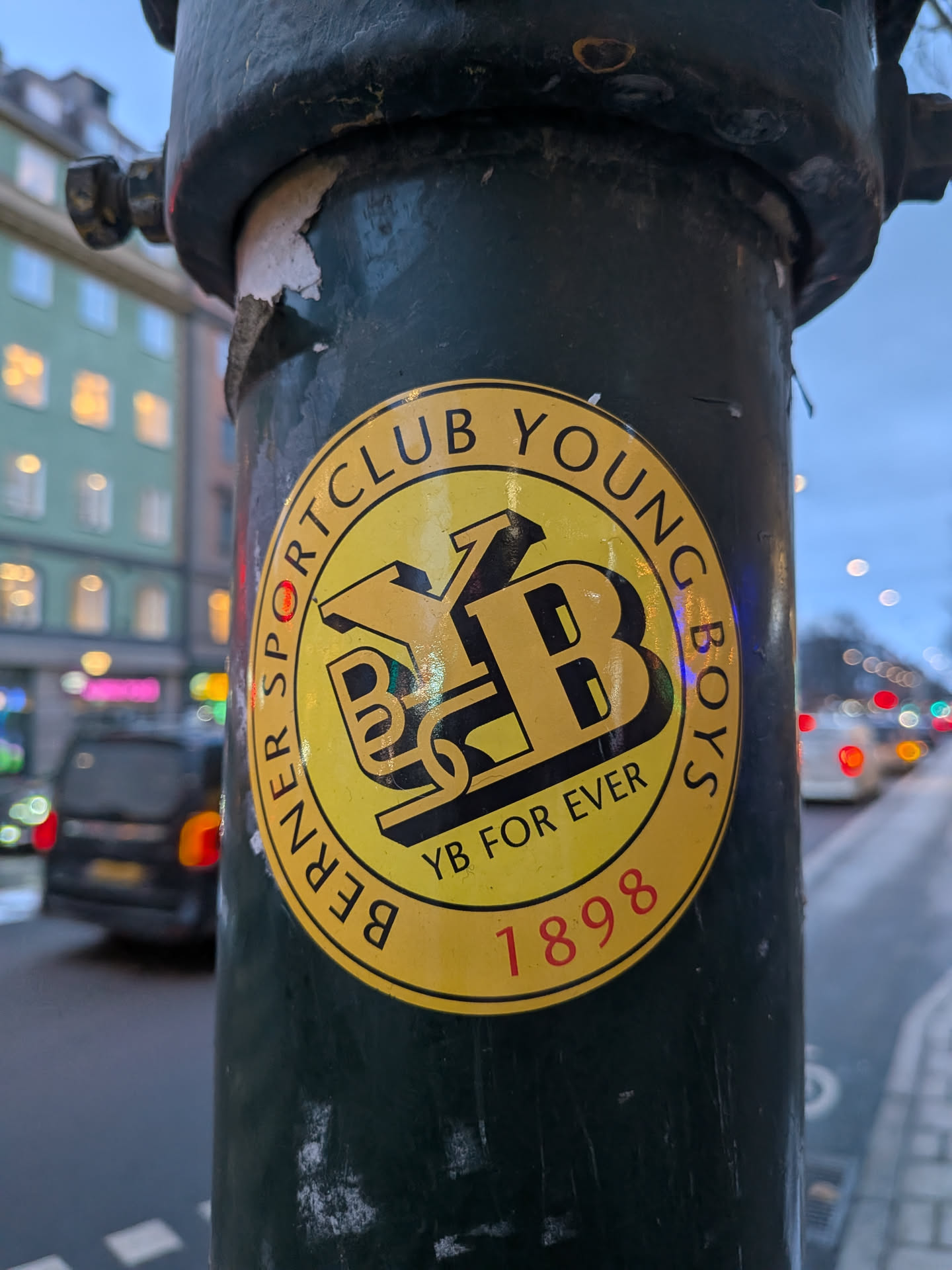 The sticker is circular, featuring a vibrant yellow background with black text and a central black emblem. An outer ring contains the words "BERNER SPORTCLUB YOUNG BOYS" in black capital letters. Inside this ring, at the bottom, the phrase "YB FOR EVER" is written in black, with the year "1898" in red numbers directly below it. The center of the sticker showcases a prominent, stylized, three-dimensional 'YB' monogram in black, designed with a shadow effect to give it depth. The sticker appears slightly worn, indicating it's a physical object adhered to a dark green pole.