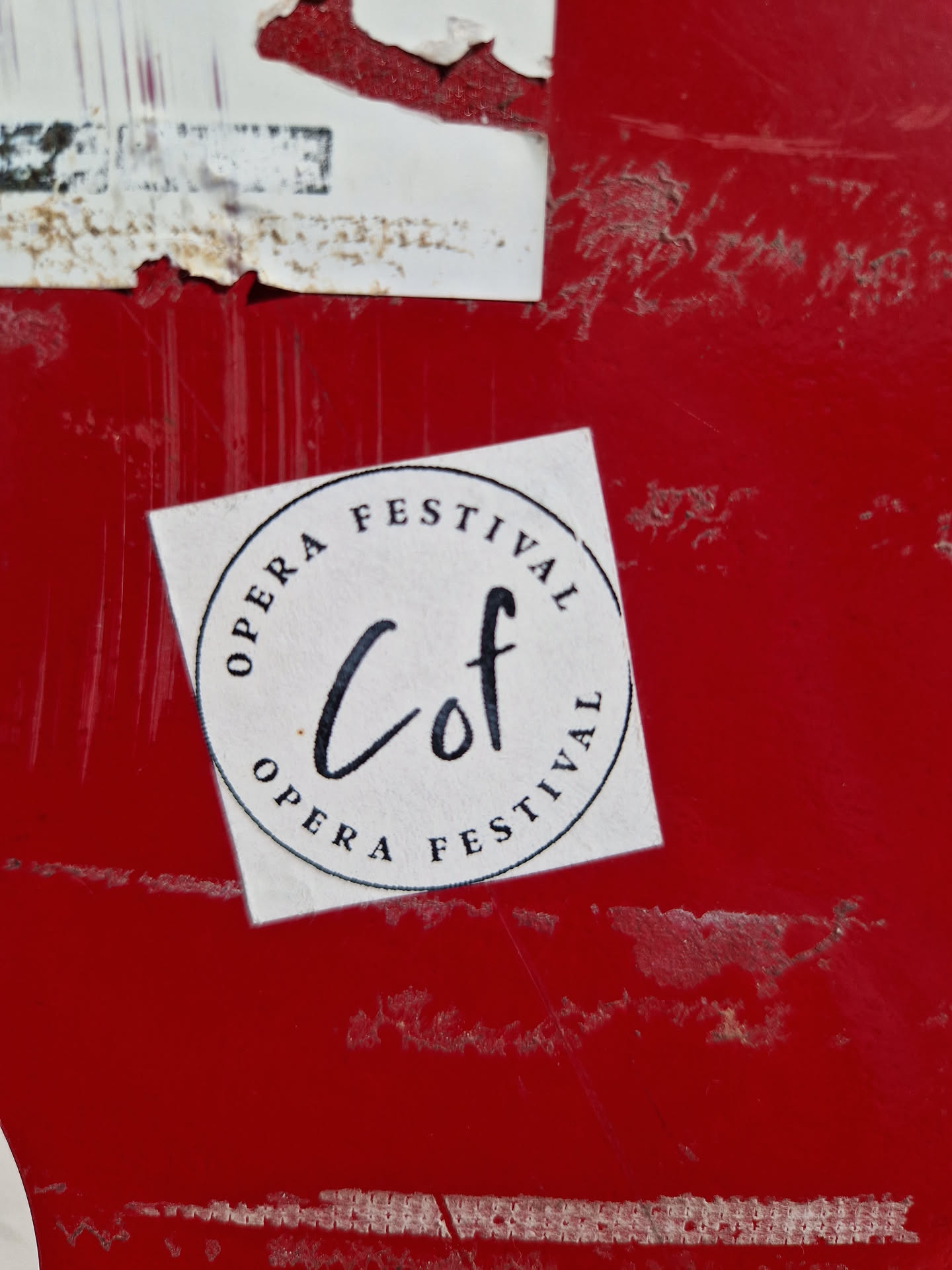 A round white sticker with black text on a red surface. The text in the circle says "Opera Festival" twice, with the abbreviation "Cof" written in a stylized script in the center.