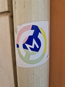 A round sticker with a stylized letter 'M' in a blue segment, set against a background of pastel pink, yellow, and green sections. The overall style is geometric and minimalist.