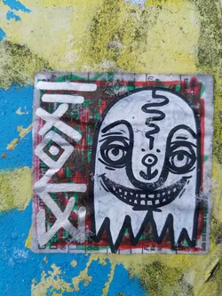 A sticker featuring a whimsical ghost character with a wide grin. The design is predominantly black and white with a touch of red and green visible from the underlying sticker. It has been stuck to a blue and yellow painted surface, adding a pop of art to the urban environment.