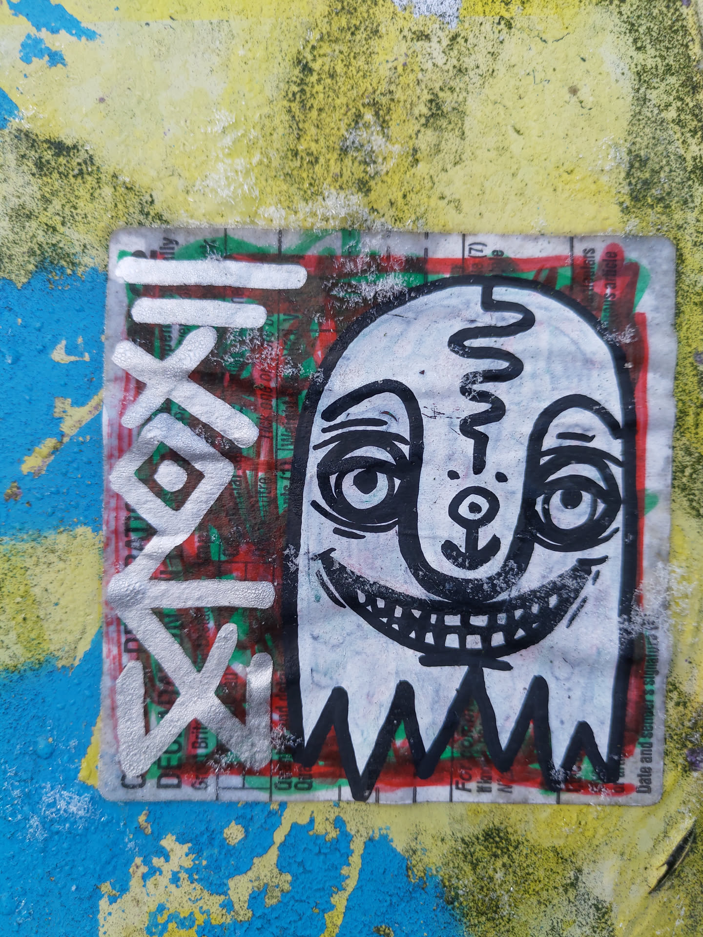 A sticker featuring a whimsical ghost character with a wide grin. The design is predominantly black and white with a touch of red and green visible from the underlying sticker. It has been stuck to a blue and yellow painted surface, adding a pop of art to the urban environment.